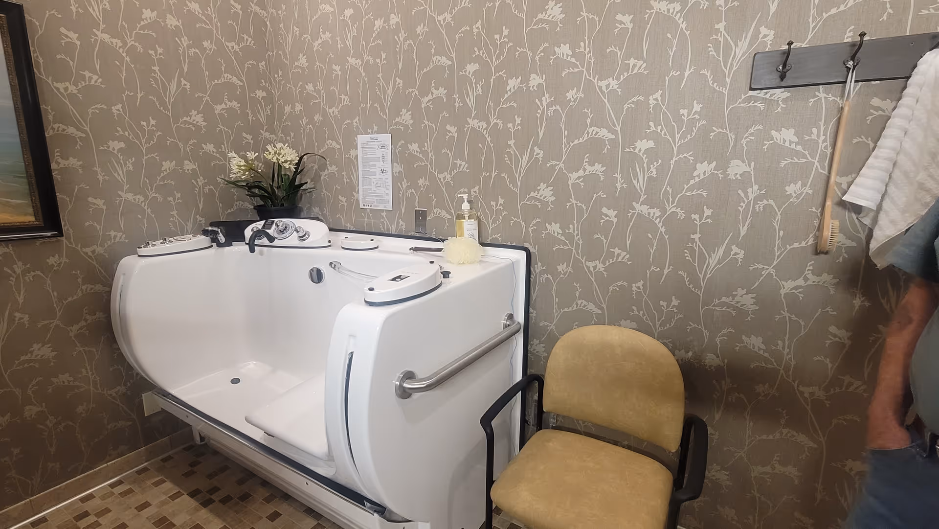 A specialized walk-in bathtub with controls and a grab bar, placed against a wall with floral patterned wallpaper. Next to the bathtub is a beige chair with black armrests. A towel rack with hanging towels and a bath brush is mounted on the wall. A small plant and a bottle of soap are placed on the bathtub ledge.