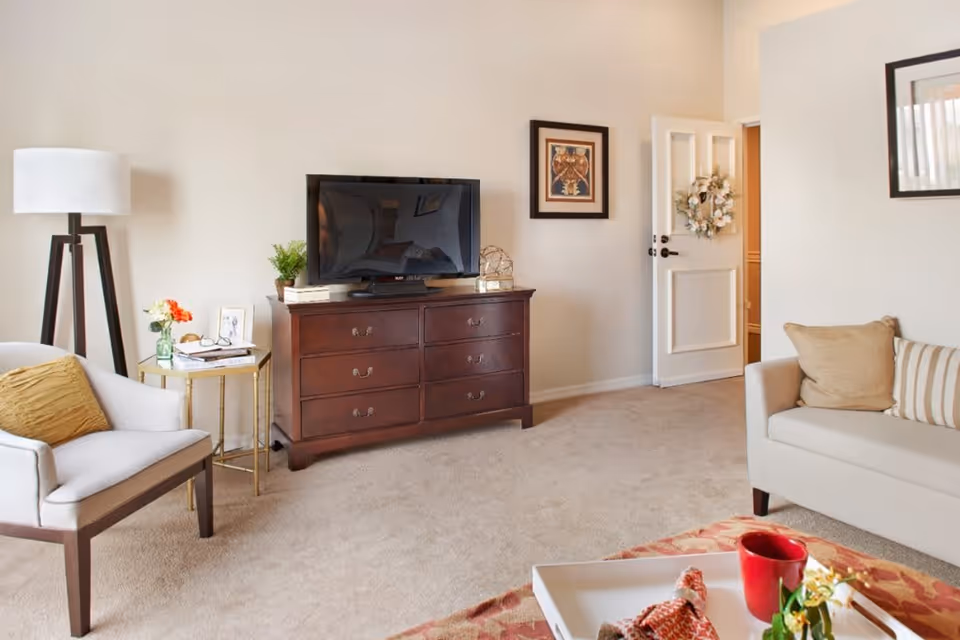 A cozy living room with beige carpet and light-colored walls. There is a wooden dresser with a flat-screen TV on top, a framed picture on the wall, and an open door with a decorative wreath. To the left, there is a white armchair with a mustard-yellow pillow next to a small round side table holding a vase with flowers and some books. On the right, a beige sofa with striped and solid pillows is partially visible. A white tray with a red cup and decorative items is on a patterned rug in the foreground.