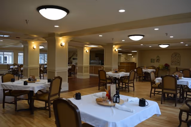 Dining room with multiple tables set with white tablecloths and chairs beneath warm ceiling lights and columns.