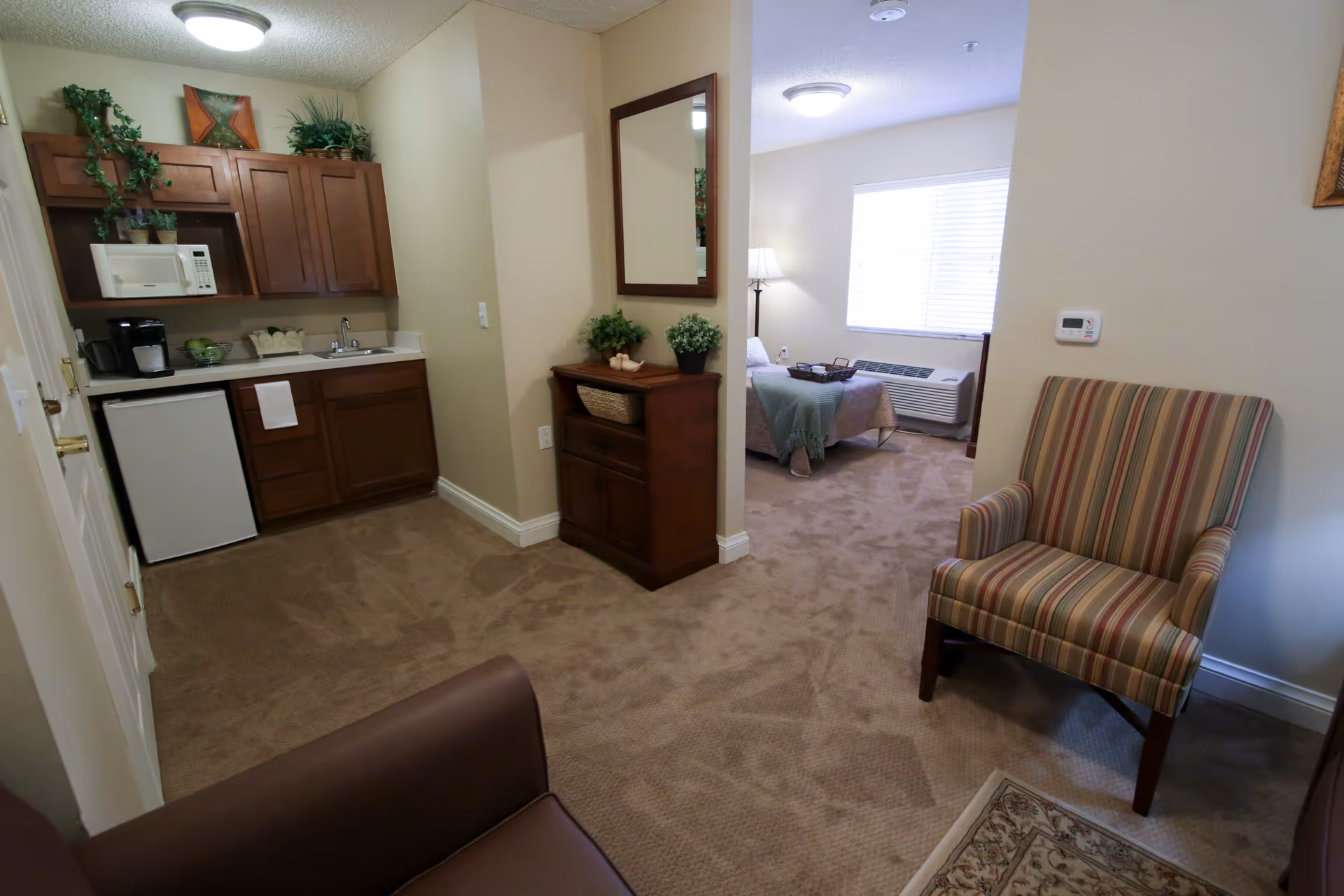 Interior view of a senior living facility apartment showing a small kitchenette with wooden cabinets, a microwave, coffee maker, and mini fridge on the left. A hallway leads to a bedroom with a bed, lamp, and window with blinds. In the foreground, there is a striped armchair and part of a brown sofa on a carpeted floor. A wooden cabinet with plants and a mirror is positioned between the kitchenette and bedroom.