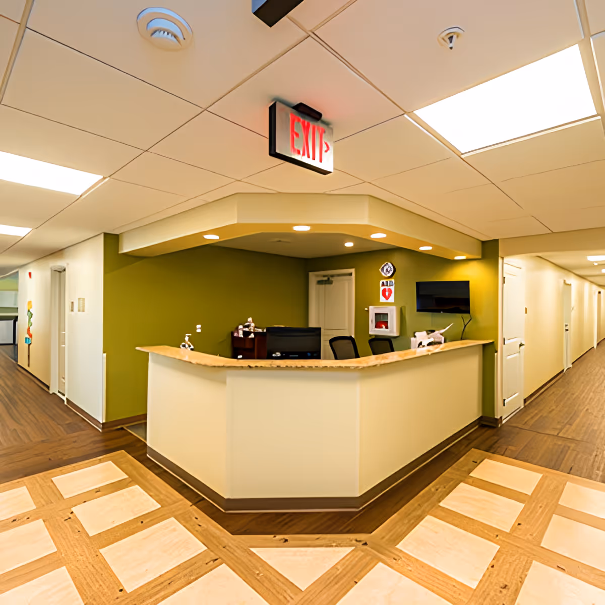 Reception desk area in a senior living community with a green accent wall, overhead exit sign, computer monitors, chairs, and a hallway extending on both sides.