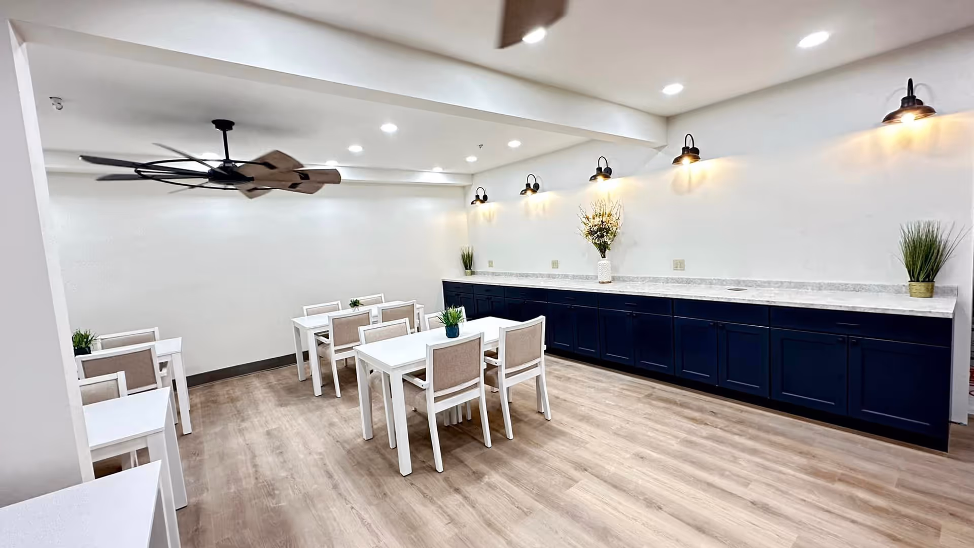 A clean, modern dining area with several white tables and beige cushioned chairs arranged neatly on a light wood floor. The room features a long countertop with dark blue cabinets underneath, decorated with small plants and a vase of flowers. Five wall-mounted lights illuminate the space, and a large ceiling fan is visible above.