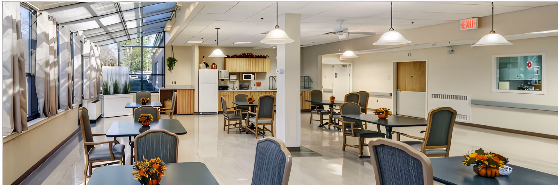 Bright communal dining area with several tables and chairs, large windows/skylight along the left, and a small kitchen/service area at the back.