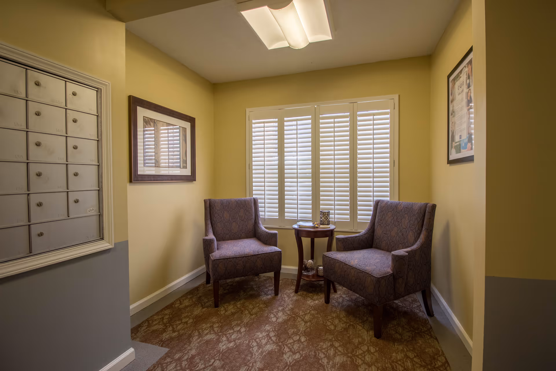 Two upholstered chairs and a small round table in a quiet seating nook with shuttered windows and mailboxes on the wall.