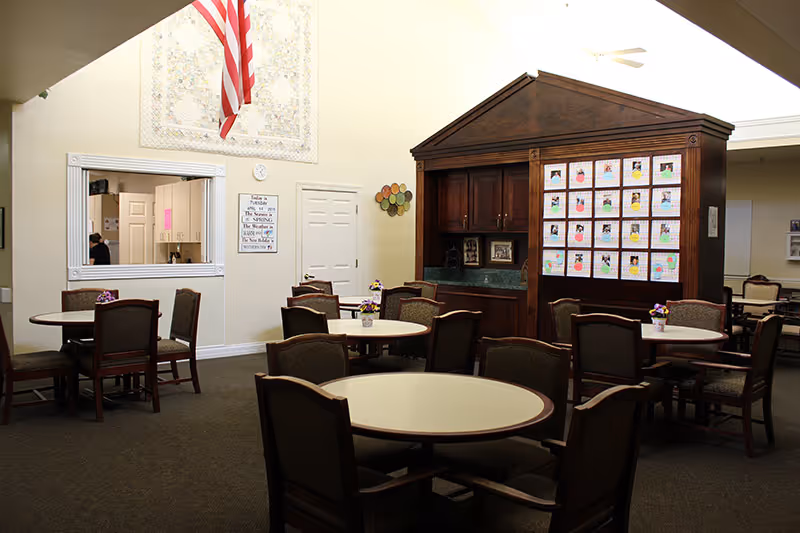 A common area with multiple round tables and chairs arranged for seating. There is a wooden cabinet with colorful drawings or artwork displayed on it. An American flag hangs on the wall above a service window and a door. The room has a carpeted floor and neutral-colored walls.