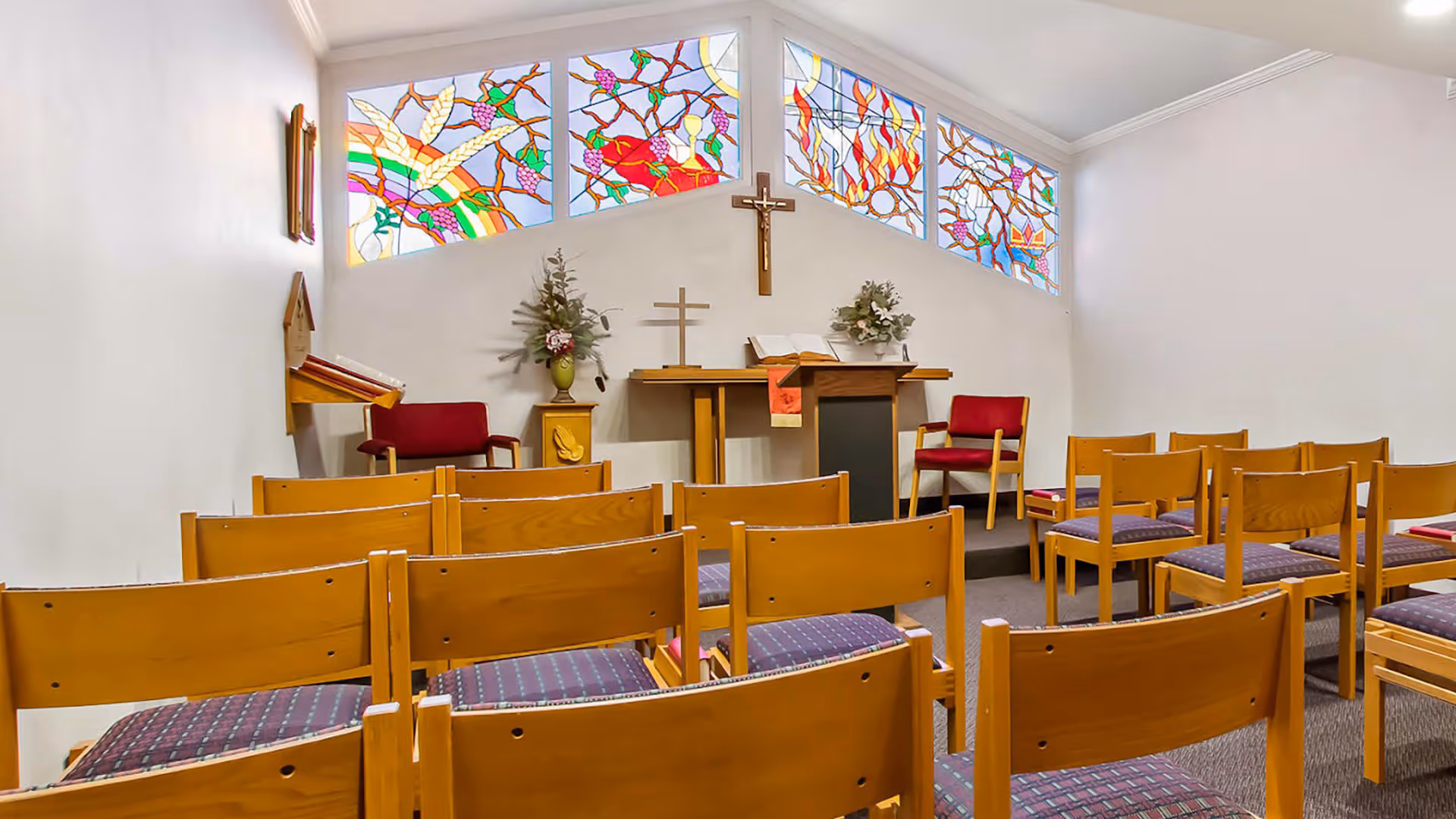 Interior view of a small chapel or prayer room with wooden chairs arranged in rows facing a wooden altar. The altar has two crosses, an open book, and floral arrangements. Above the altar are colorful stained glass windows depicting various designs. There are two red cushioned chairs on either side of the altar.