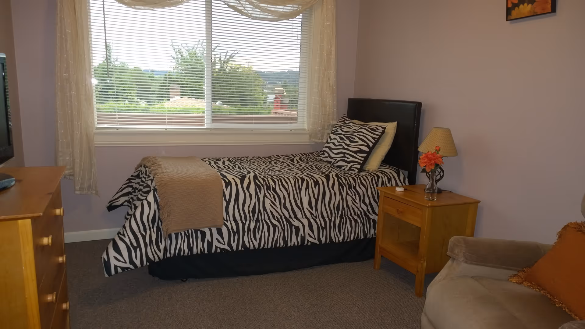 A small bedroom with a single bed covered in zebra print bedding and a beige throw blanket. Next to the bed is a wooden nightstand with a lamp and a small vase with orange flowers. There is a window with blinds and sheer curtains behind the bed, showing greenery outside. A wooden dresser with a TV on top is on the left side of the room, and a beige armchair with an orange pillow is partially visible on the right.