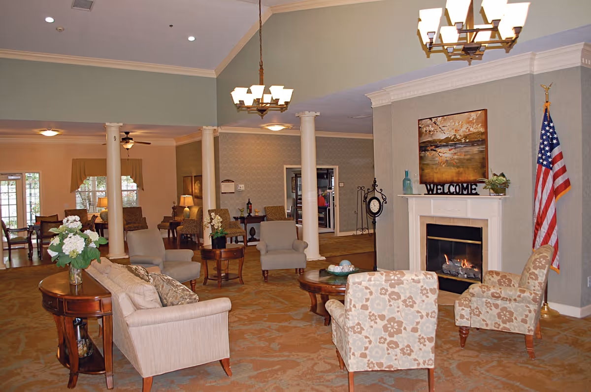 A spacious and warmly decorated living room area in a senior living facility featuring a beige sofa, patterned armchairs, wooden coffee tables, a fireplace with a 'WELCOME' sign and artwork above it, an American flag, and soft lighting from chandeliers and lamps.