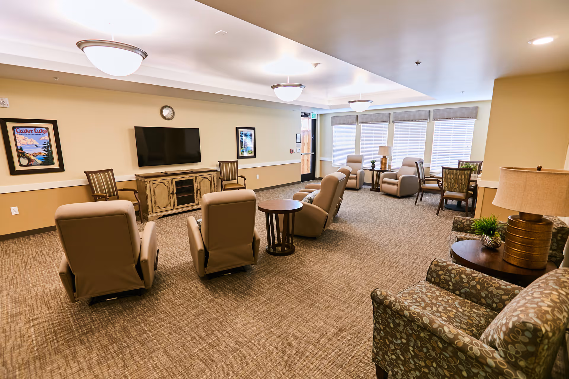 A spacious senior living common area with several beige recliner chairs arranged facing a wall-mounted flat screen TV. The room has carpeted floors, beige walls, and large windows with blinds letting in natural light. There are side tables with lamps and plants, framed artwork on the walls, and additional seating including patterned armchairs and wooden chairs around small tables.