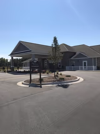 Exterior view of a single-story building with a covered entrance and a small landscaped island with a tree and plants in front. A sign near the entrance reads 'Vicinia Intermediate'. The sky is clear and blue.