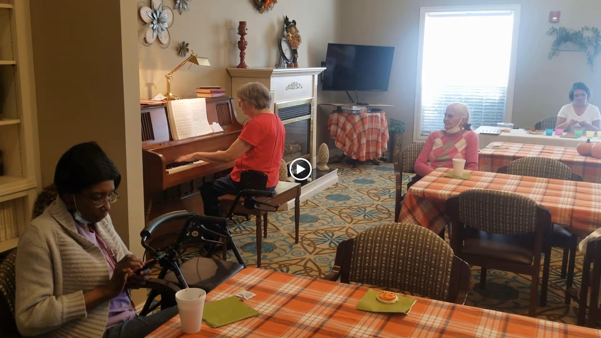 Several residents sit at plaid-covered dining tables while someone plays a piano in a communal activity room.