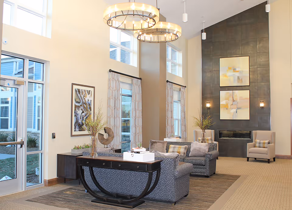 A bright and spacious living room area in a senior living facility with high ceilings and large windows. The room features two patterned sofas with cushions, a dark wooden console table with decorative items, two armchairs with plaid pillows, a modern fireplace with two abstract paintings above it, and two circular chandeliers hanging from the ceiling. Light-colored curtains frame the windows, and the floor is carpeted with a patterned rug under the seating area.