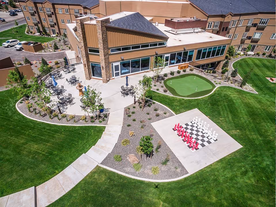 Aerial view of an outdoor area at a senior living facility showing a patio with tables and chairs, a small putting green, a large outdoor chessboard with oversized red and white chess pieces, surrounded by well-maintained green lawns and landscaping.