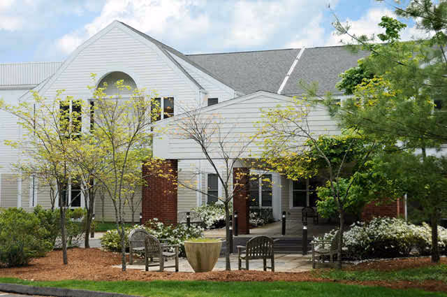 Exterior view of a senior living facility building with white siding and a gray roof, surrounded by small trees and shrubs. There is a paved area with wooden chairs and a large planter in front of the entrance.