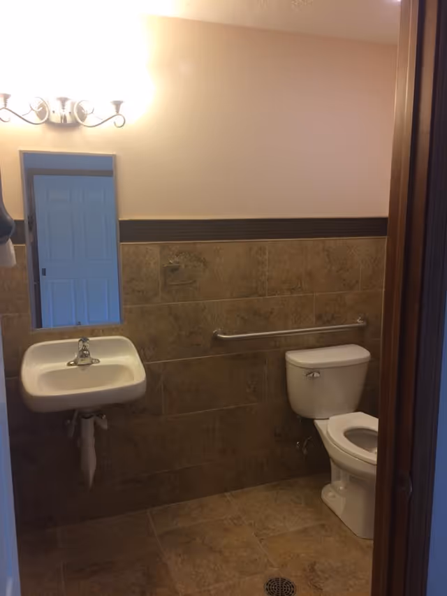 A small bathroom with beige tiled walls and floor, featuring a white toilet with a grab bar on the wall beside it, a white wall-mounted sink with a faucet, a rectangular mirror above the sink, and a light fixture with two bulbs above the mirror.