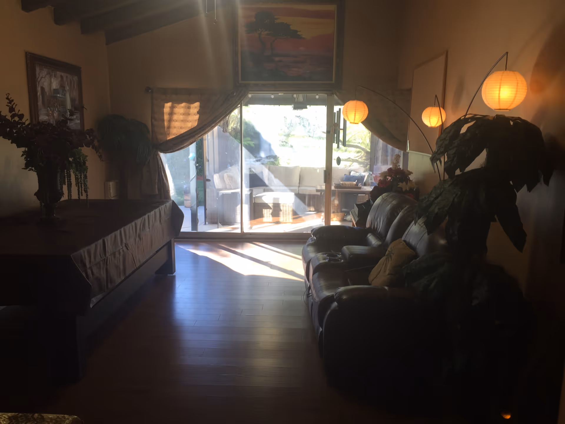A dimly lit living room with a dark leather sofa on the right side, decorated with pillows and a large leafy plant beside it. There are three hanging round paper lanterns providing warm light. On the left side, there is a table covered with a dark cloth and a large floral arrangement. The room has wooden flooring and a sliding glass door at the back leading to a bright outdoor patio area with seating. Curtains frame the sliding door, and a framed painting hangs above it.