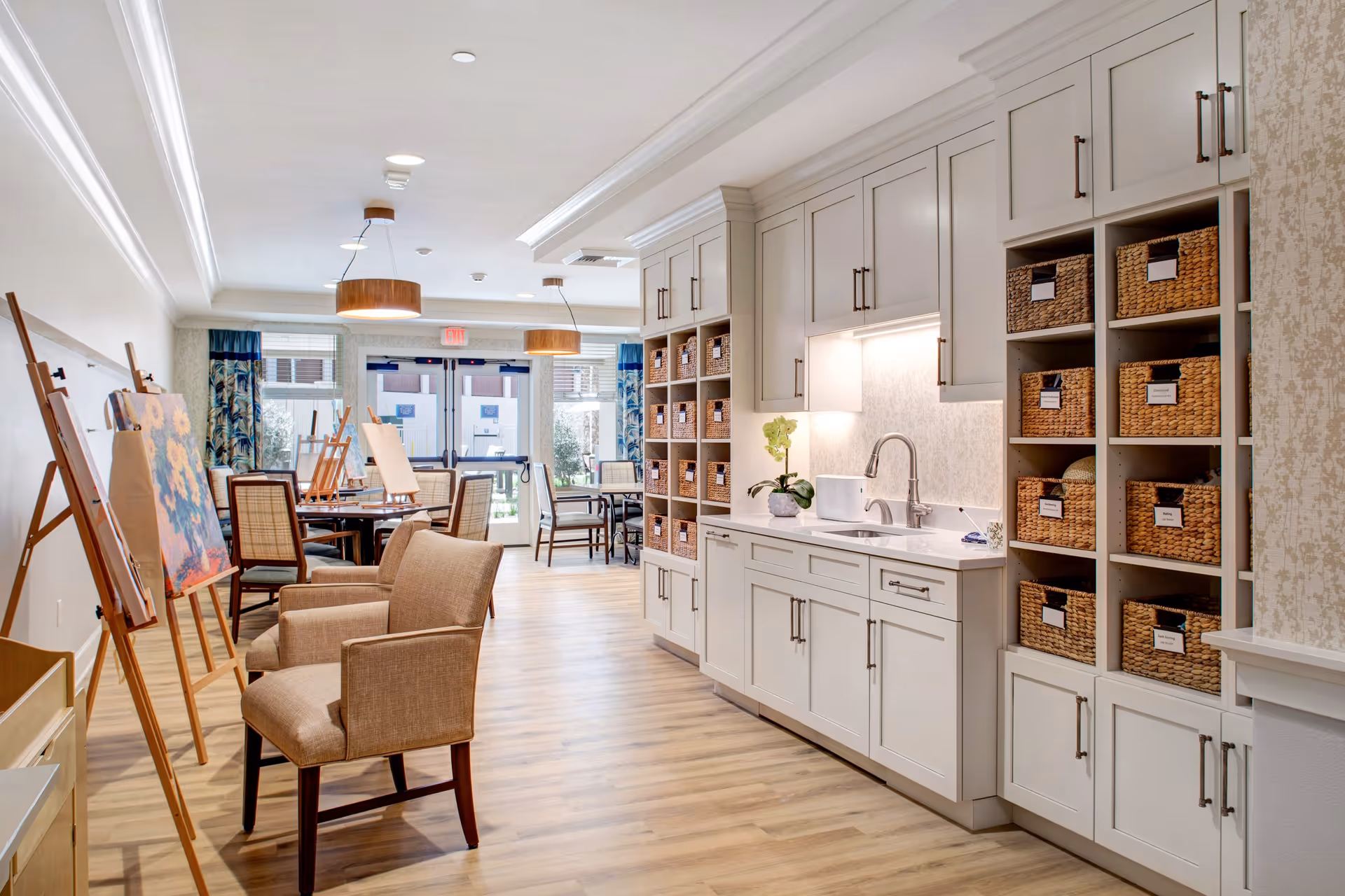 Bright and spacious art activity room in a senior living facility with wooden flooring, beige cushioned chairs, easels displaying paintings, and a white cabinetry area with a sink and woven baskets for storage. Large windows and glass doors allow natural light to enter the room.