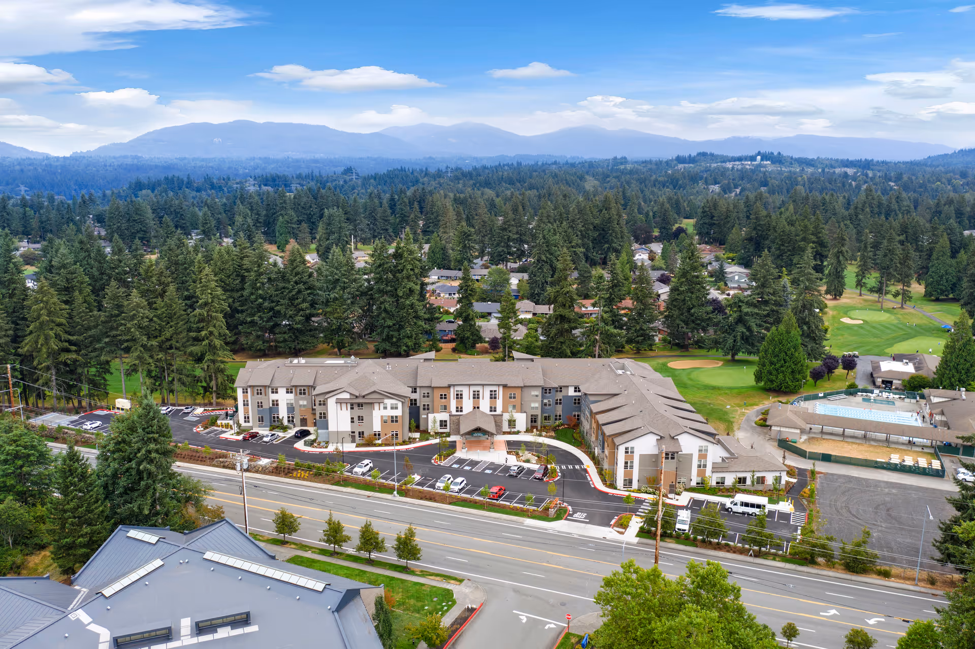 Aerial view of Village Concepts of Fairwood senior living facility surrounded by trees and greenery, with a parking lot in front and a golf course visible in the background under a partly cloudy sky.