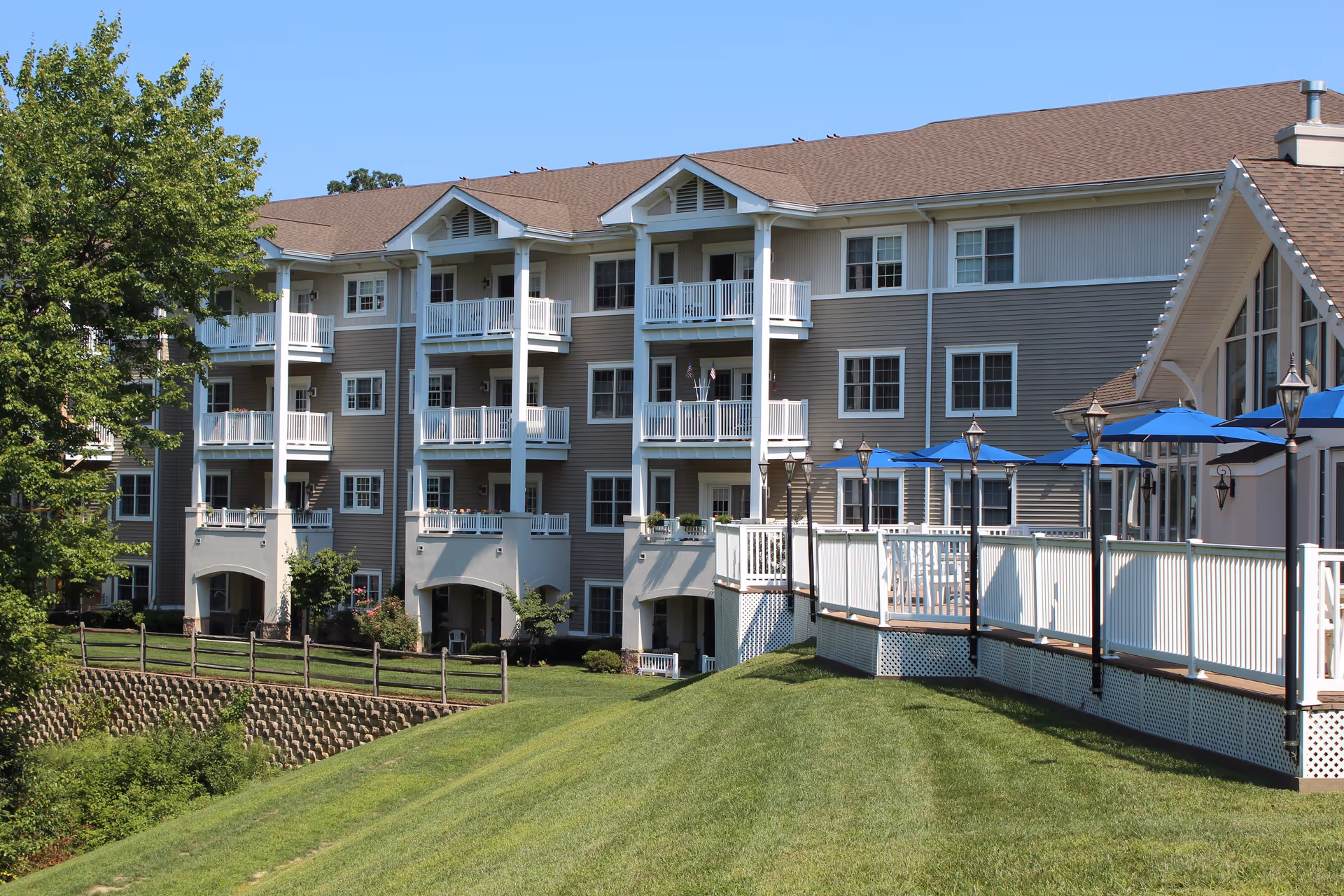 Exterior view of a multi-story senior living facility building with balconies, a well-maintained grassy lawn, trees, and a patio area with blue umbrellas and white railings under a clear blue sky.