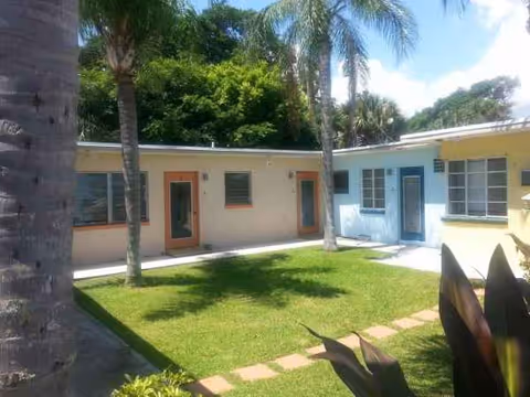 Single-story pastel facility exterior with doors, windows, palm trees, and a grassy courtyard.