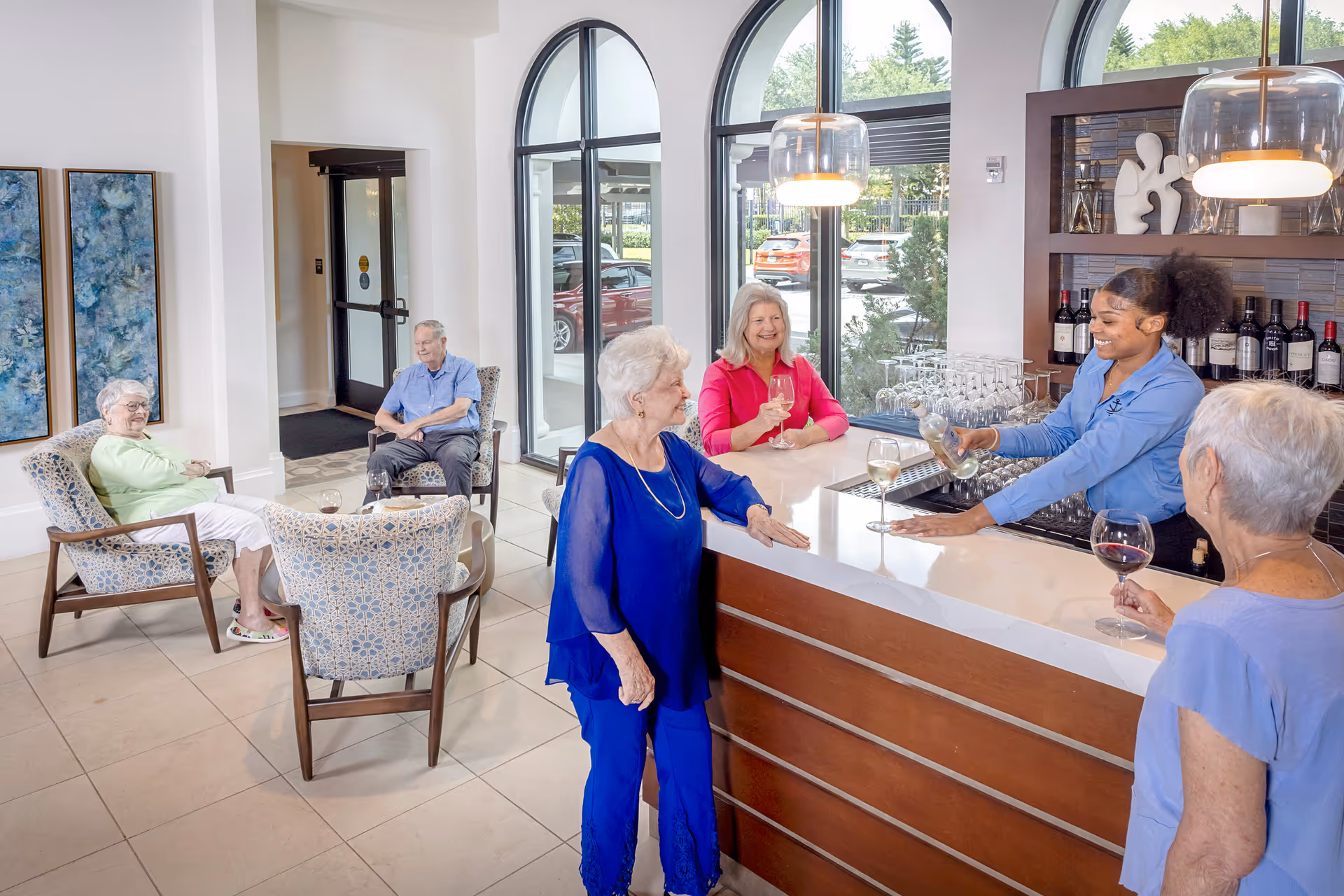 Residents gather in a bright interior lounge and bar area of a senior living facility while a staff member pours wine and others sit and chat.