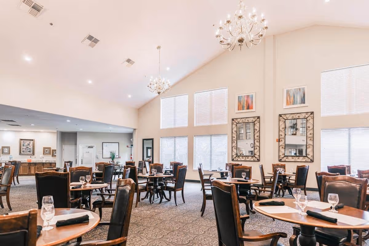 A spacious dining room with multiple round wooden tables and dark cushioned chairs. Each table is set with glasses, napkins, and placemats. The room features high vaulted ceilings with two chandeliers, large windows with blinds, decorative mirrors, and framed artwork on the walls. The far end of the room has a counter area with cabinets and additional seating.