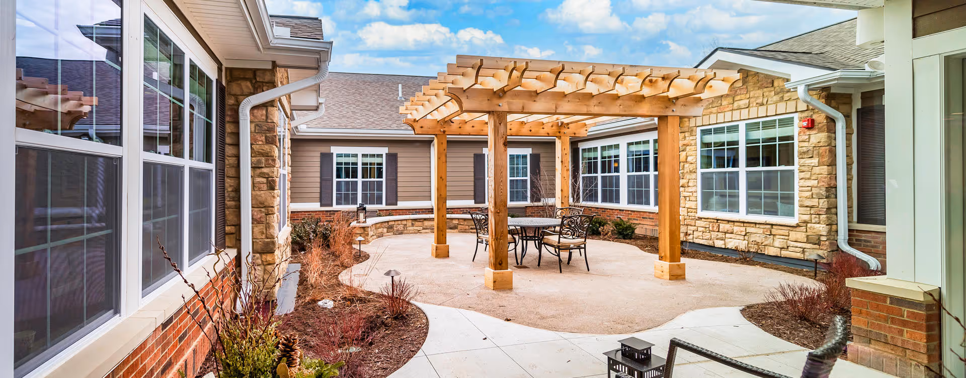 Outdoor courtyard area at Bickford of Canton featuring a wooden pergola with a round metal table and chairs underneath. The courtyard is surrounded by building walls with large windows and stone and brick accents. There are landscaped garden beds with plants and a clear blue sky with some clouds above.