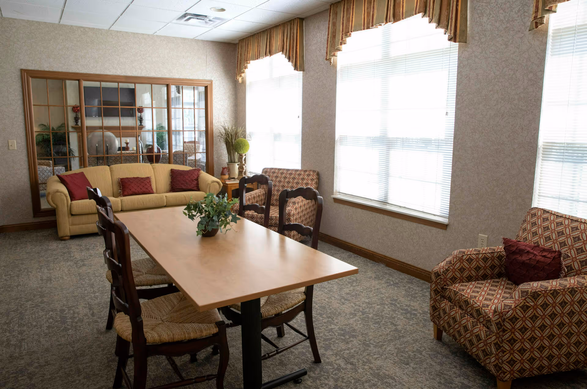 A cozy sitting area with a wooden table surrounded by four chairs, a yellow sofa with red pillows, and two patterned armchairs with red pillows. Large windows with blinds and valances allow natural light to fill the room. A large mirror with wooden framing is mounted on the wall behind the sofa, reflecting a fireplace and TV in another room.