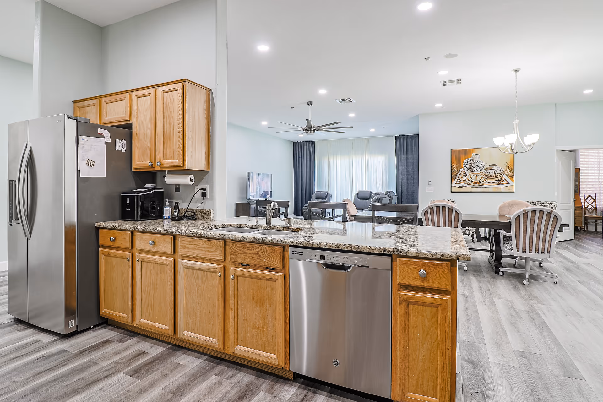 Open-plan kitchen with a granite island, stainless steel refrigerator and dishwasher, wooden cabinets, and a dining/living area visible in the background.