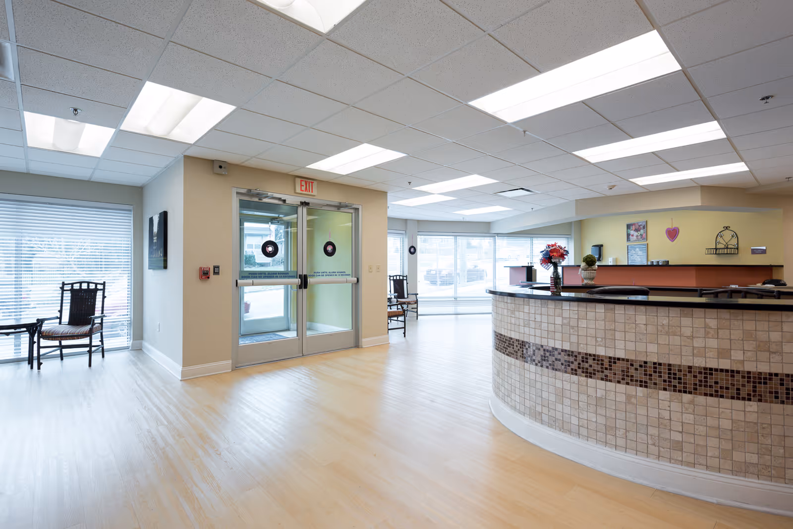 Bright and spacious reception area with a curved tiled front desk, two wooden chairs with cushions near large windows with blinds, double glass entrance doors with exit sign above, and ceiling lights illuminating the light wood flooring.