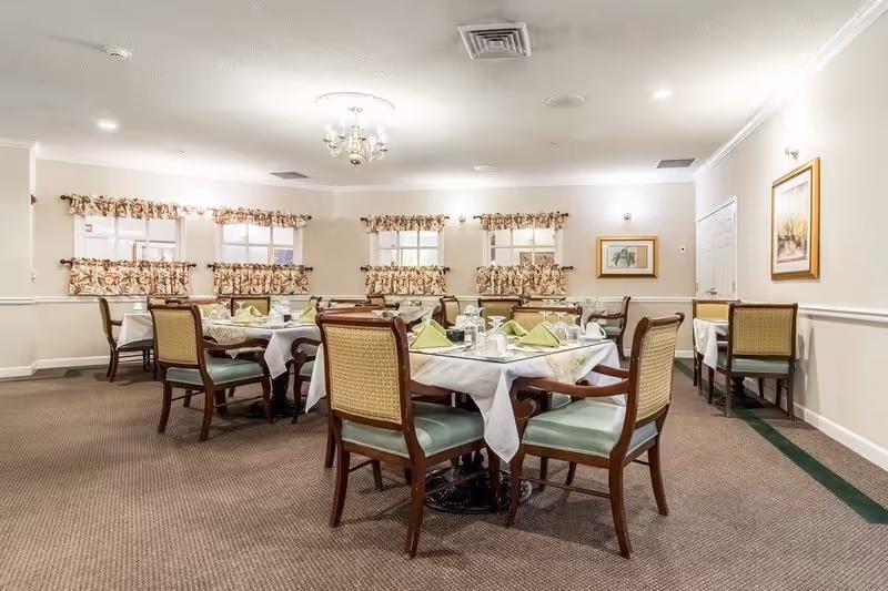 A dining room with multiple tables covered with white tablecloths and set with plates, glasses, and folded green napkins. The room has beige walls, floral curtains on the windows, framed artwork on the walls, and carpeted flooring. Wooden chairs with green cushions surround the tables.