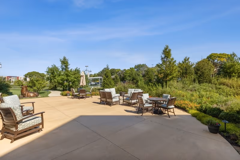 Outdoor patio area with several cushioned chairs and tables arranged on a concrete surface, surrounded by greenery and trees under a clear blue sky.