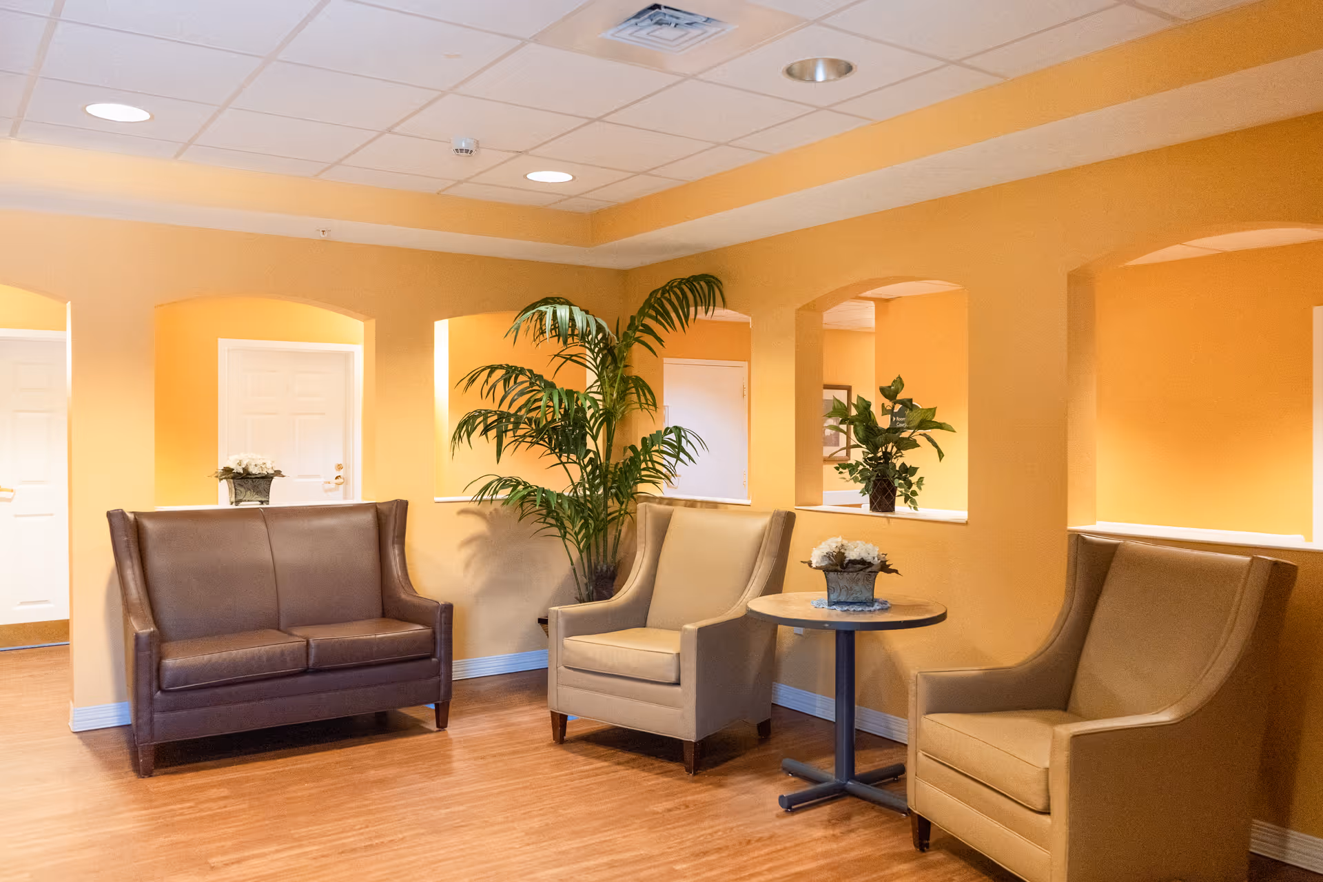 A cozy seating area in a senior living facility with two beige armchairs and one brown loveseat arranged around a small round table with a flower pot. The walls are painted a warm yellow, and there are decorative plants placed in wall niches. The floor is wooden, and the ceiling has recessed lighting.