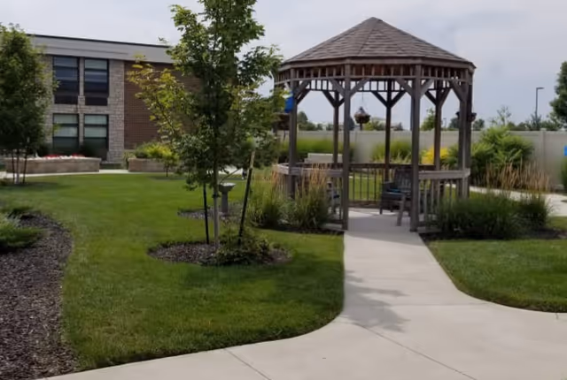 Outdoor courtyard featuring a wooden gazebo, paved walkway, green lawn, and a brick building in the background.