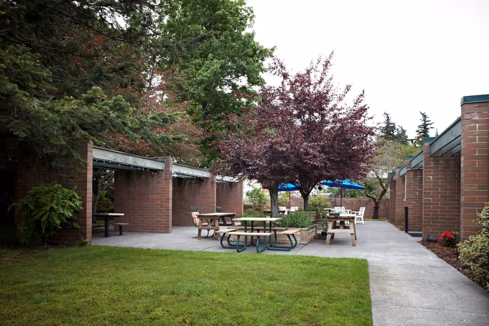 Outdoor patio area with multiple picnic tables and benches surrounded by brick walls and green trees, including a tree with purple leaves. There are blue umbrellas in the background providing shade over some tables. The ground is a mix of concrete and grass.