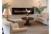 Seating area with upholstered chairs and a loveseat arranged around a wooden coffee table in front of a wall-mounted electric fireplace, flanked by potted plants and a table lamp.