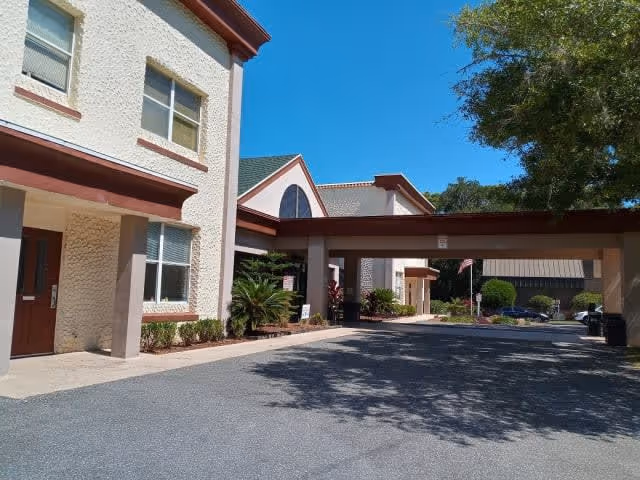 Exterior view of a senior living facility building with beige textured walls and brown trim under a clear blue sky. The building has multiple windows and a covered driveway or entrance area. There are some small bushes and plants near the building, and a large tree casting shadows on the pavement.