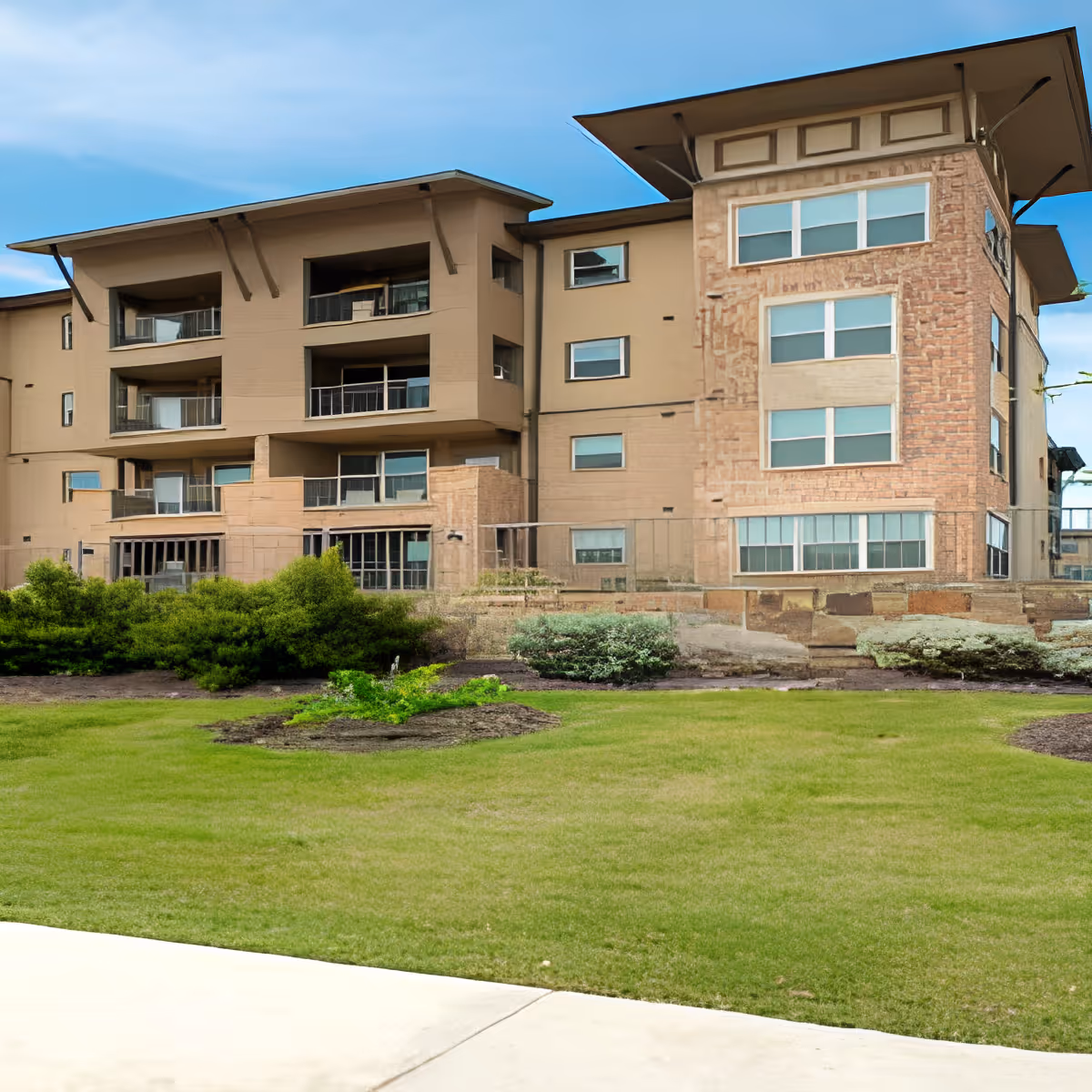 Exterior view of a multi-story senior living facility building with beige and stone facade, multiple windows, balconies, and a well-maintained lawn with shrubs in the foreground under a blue sky.