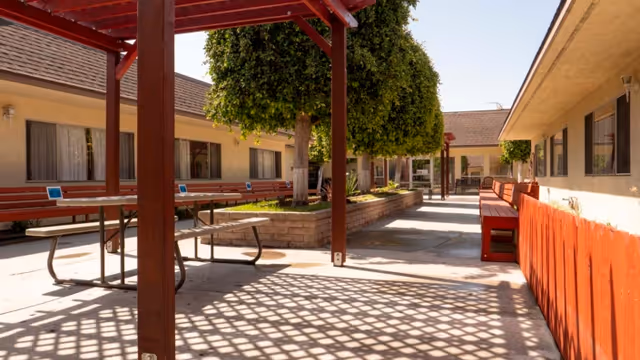 Outdoor courtyard area with shaded pergolas, picnic tables, benches, and neatly trimmed trees surrounded by single-story building walls with windows.