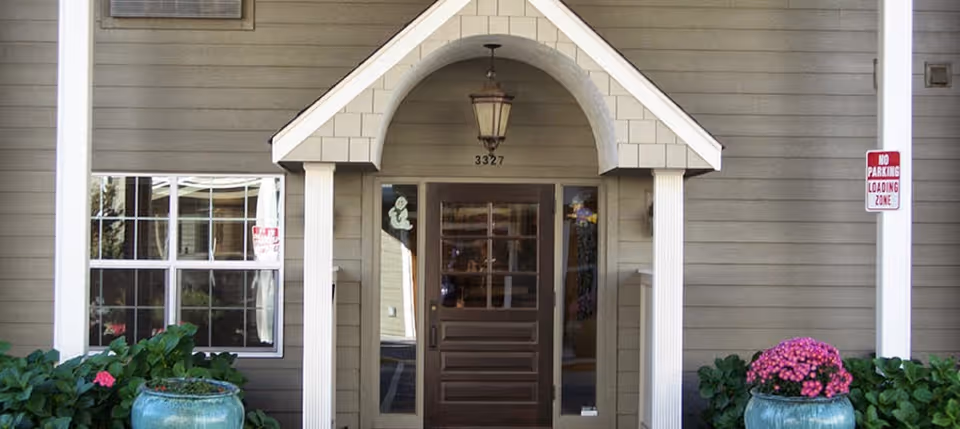 Covered main entrance with a wooden door and transom under an arched portico, flanked by windows and potted plants.