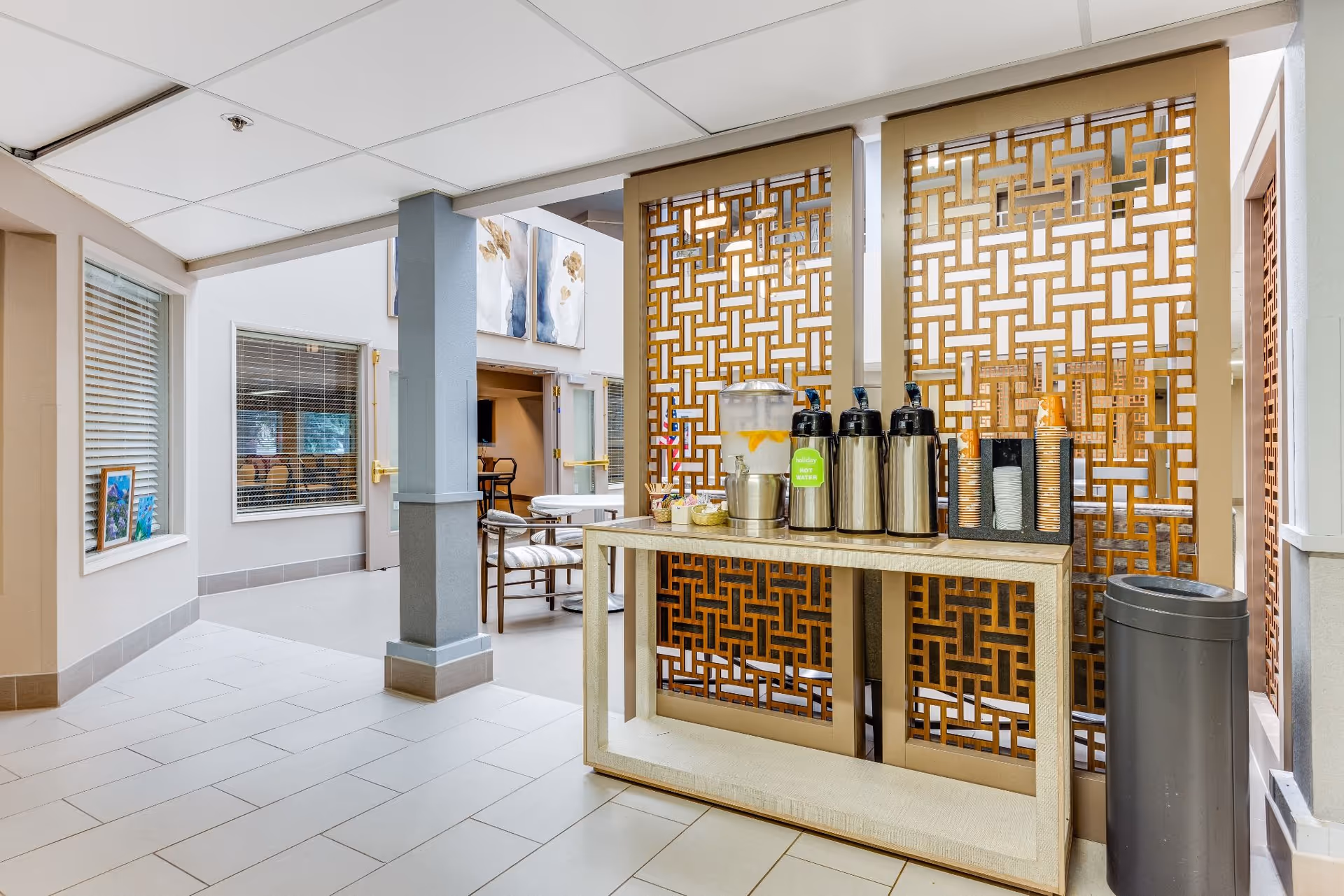 A beverage station with a water dispenser containing lemon water, three stainless steel coffee dispensers, and a holder with disposable cups, set against decorative wooden lattice panels in a bright, tiled hallway area with seating and artwork visible in the background.