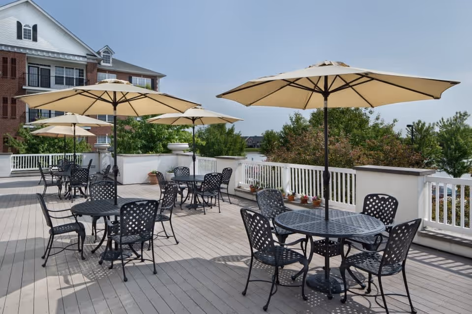 Outdoor patio area with multiple black metal tables and chairs, each table shaded by a large beige umbrella. The patio has a light wooden floor and white railing, with greenery and a building visible in the background under a clear blue sky.