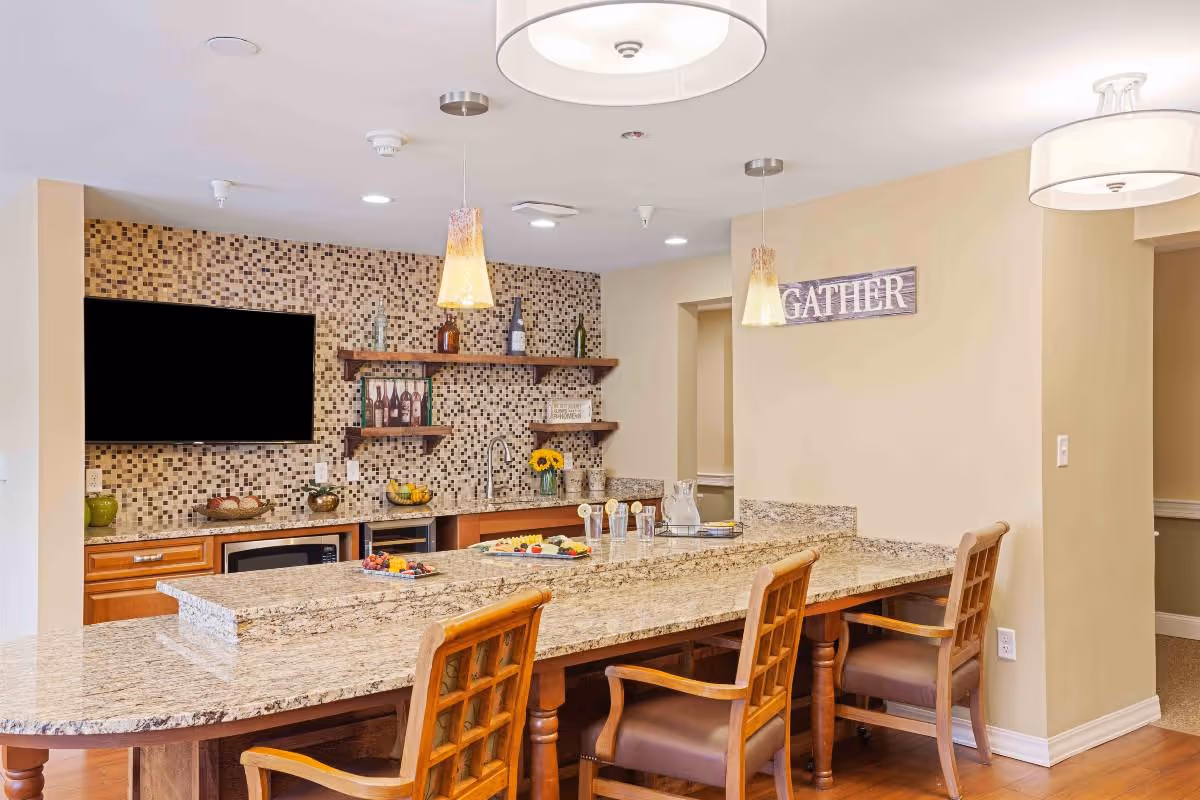 A modern kitchen area with a large granite countertop island surrounded by wooden chairs with armrests. The back wall features a mosaic tile backsplash, open wooden shelves with decorative bottles and items, a mounted flat-screen TV, and a sink. Two pendant lights hang over the island, and a sign on the wall reads 'GATHER'. The room has warm lighting and a cozy atmosphere.