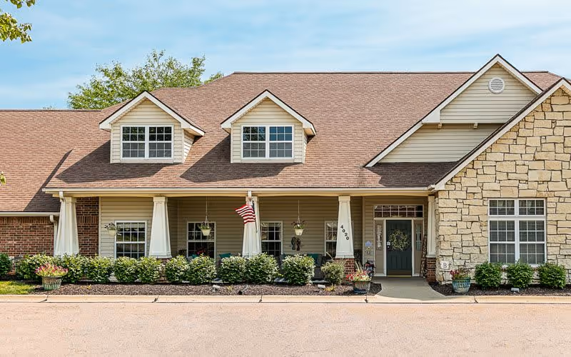 Front exterior view of a single-story senior living facility building with a brown shingled roof, beige siding, stone accents, and a covered porch with white columns. There are several windows, hanging plants, an American flag, and neatly trimmed bushes along the front. The entrance door is dark green with a wreath.