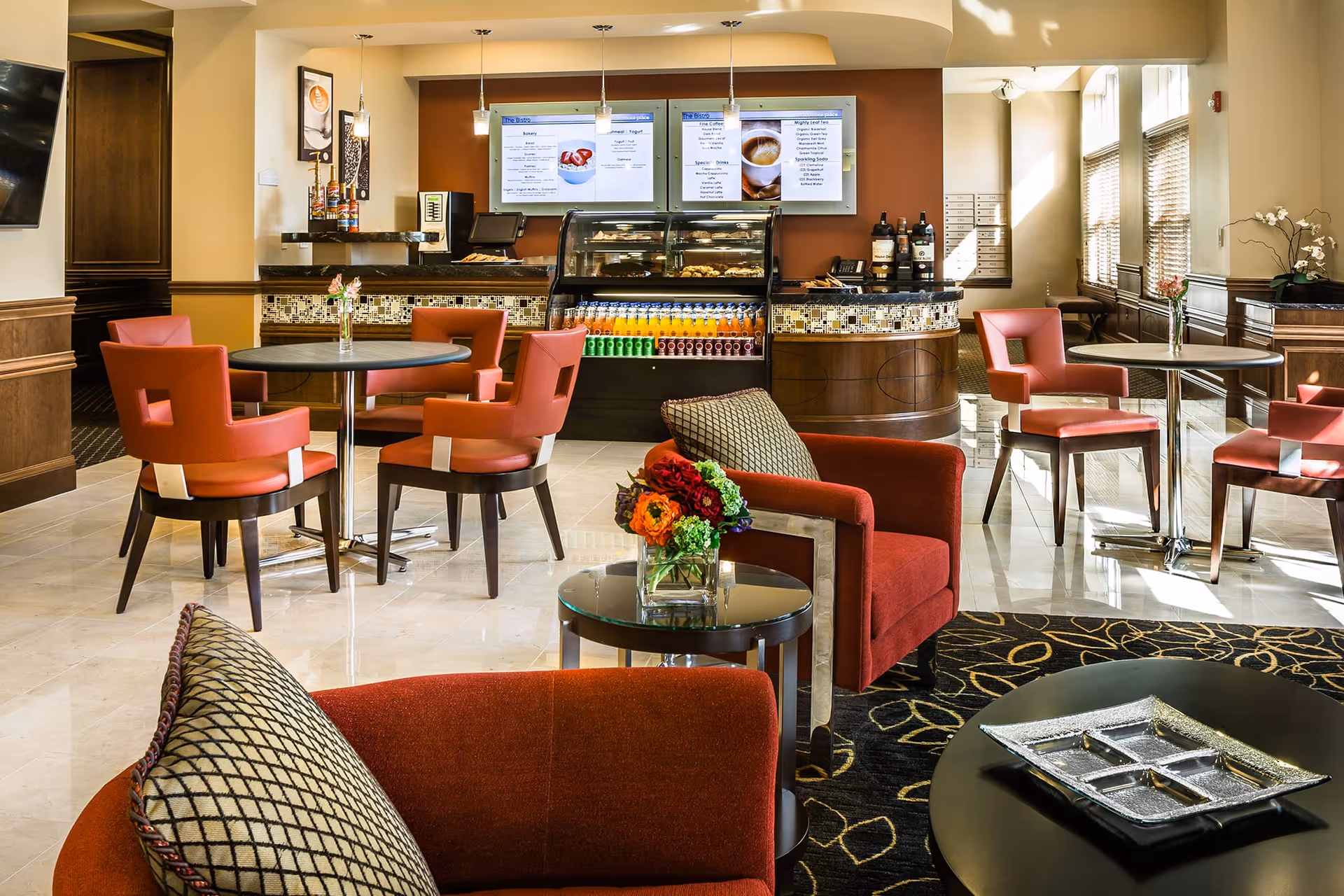 A bright and modern common area with red cushioned chairs and round tables. In the background, there is a counter with a display case filled with beverages and snacks, and two digital menu boards mounted on the wall. The room has large windows allowing natural light to fill the space, and decorative flowers are placed on the tables.