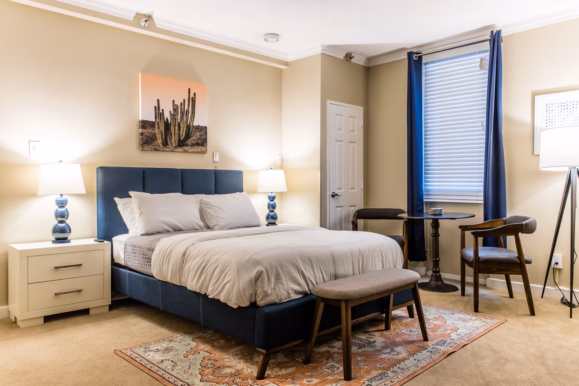 A neatly made bed with white linens and a blue upholstered headboard in a bedroom. There are two bedside tables with blue lamps on each side of the bed. A small bench is placed at the foot of the bed on a patterned rug. To the right, there is a round table with two wooden chairs near a window with blue curtains and white blinds. A floor lamp stands in the corner, and a cactus picture hangs on the wall above the bed.