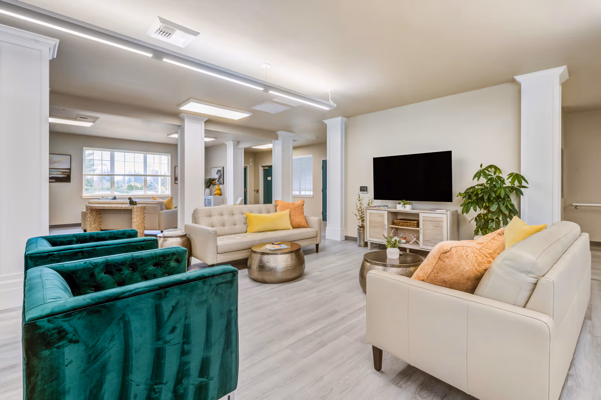A bright and spacious senior living facility common area with modern decor. The room features two beige sofas with orange and yellow pillows, two green velvet armchairs, two round metallic coffee tables, a large flat-screen TV mounted on the wall above a light wood cabinet, and a large window letting in natural light. There are white columns and light wood flooring throughout the space.