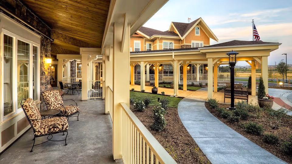 Covered outdoor walkway with cushioned chairs and tables along the side of a senior living facility building, leading to a landscaped garden area with a paved path, benches, lamp posts, and an American flag in the background during early evening.