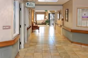 A clean, well-lit hallway in a senior living facility with tiled floors, beige walls, handrails on both sides, and a few chairs placed along the corridor. There is a sign overhead indicating the direction to the Day Room and Therapy, and a window at the far end letting in natural light.