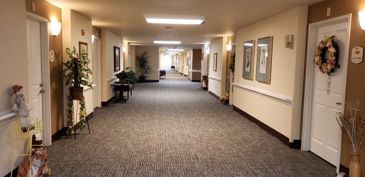 A long, well-lit hallway in a senior living facility with beige walls and carpeted floor. The hallway has several doors on both sides, some decorated with wreaths and nameplates. There are plants and framed pictures along the walls, with benches and small tables placed intermittently. The lighting is provided by ceiling fixtures and wall sconces.